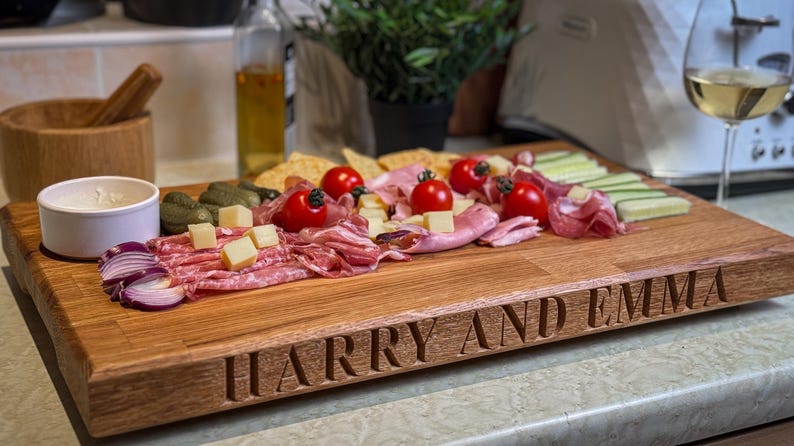 Puede incluir: Una tabla de servir de madera con los nombres "Harry y Emma" grabados en la parte delantera. La tabla est&aacute; llena de una variedad de alimentos, incluyendo salami, queso, uvas rojas y pepinos.