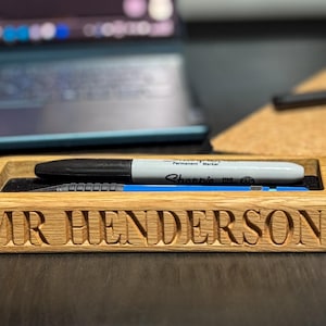 May include: Wooden desk tidy engraved with "MR HENDERSON". It contains a black Sharpie marker and a blue mechanical pencil. The desk tidy sits on a dark surface. A laptop and notebook are in the background.