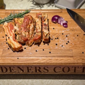 Puede incluir: Una tabla de cortar de madera con el texto "GARDENERS COTTAGE" grabado en la parte delantera. La tabla es de color marr&oacute;n claro y est&aacute; hecha de madera de roble. Hay varios trozos de panceta sazonada en la tabla, junto con una ramita de romero, algunos granos de pimienta y una cebolla roja en rodajas.