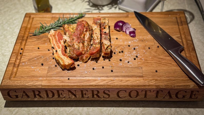 Puede incluir: Una tabla de cortar de madera con el texto "GARDENERS COTTAGE" grabado en la parte delantera. La tabla es de color marr&oacute;n claro y est&aacute; hecha de madera de roble. Hay varios trozos de panceta sazonada en la tabla, junto con una ramita de romero, algunos granos de pimienta y una cebolla roja en rodajas.