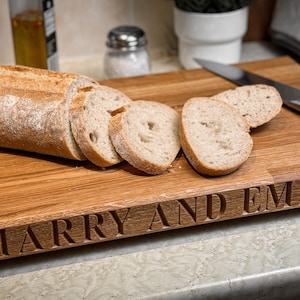 Puede incluir: Una tabla de cortar de madera con los nombres "Harry y Emma" grabados en la parte delantera. La tabla es de madera marr&oacute;n claro y tiene un acabado natural. Hay rebanadas de pan en la tabla, junto con un cuchillo de plata.
