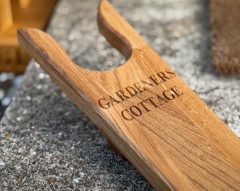 Puede incluir: Un sacabotas de madera con la inscripción "Gardeners Cottage" grabada. El sacabotas es de madera marrón clara y tiene un mango curvado. El fondo es borroso.