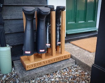 Puede incluir: Un zapatero de madera con cuatro ranuras para guardar botas de lluvia. El zapatero está hecho de madera natural y tiene las palabras "Gardeners Cottage" grabadas en la parte delantera.