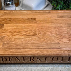 Puede incluir: Una tabla de cortar de madera grande, rectangular, con un borde elevado y un borde biselado. La tabla est&aacute; hecha de madera marr&oacute;n claro y tiene una veta de madera natural. Las palabras "GARDENERS COTTAGE" est&aacute;n talladas en el borde inferior de la tabla.