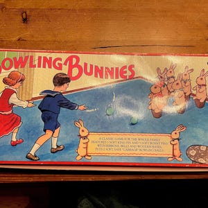 Puede incluir: Caja de juego vintage "Bowling Bunnies". La caja presenta una colorida ilustración de niños jugando a los bolos con bolos de conejitos. El texto "Bowling Bunnies" es rojo. El juego incluye bolos blandos, cintas y bases de madera.
