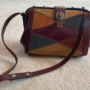 May include: A vintage patchwork leather purse with a long, adjustable strap. The bag features a geometric design in shades of brown, orange, green, and burgundy. It has a clasp closure with a decorative metal accent.
