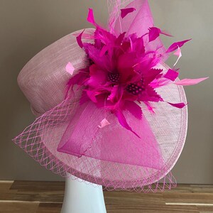 May include: A pink and fuchsia hat with a large flower and feathers. The hat is made of a straw-like material and has a net veil.