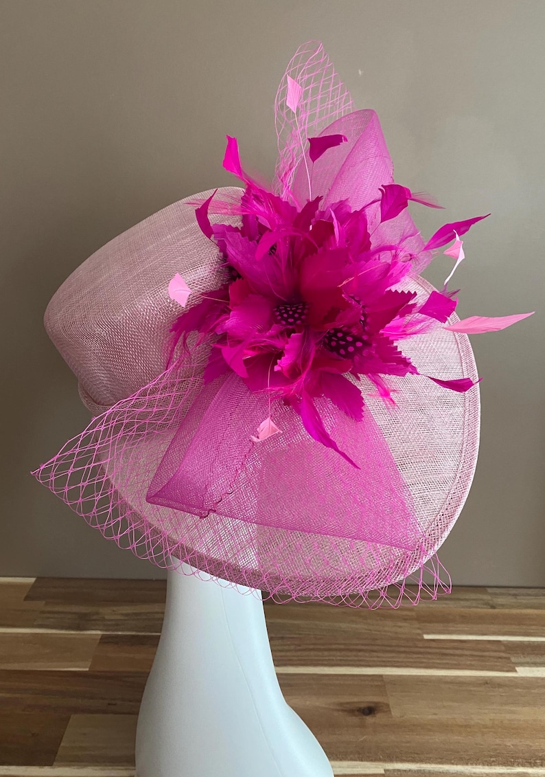 May include: A pink and fuchsia hat with a large flower and feathers. The hat is made of a straw-like material and has a net veil.