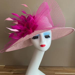 May include: A pink and fuchsia hat with a large brim and a feather accent. The hat is made of a delicate fabric and has a net overlay.