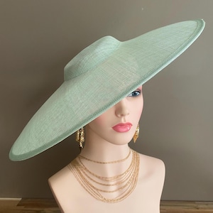 May include: A light green, wide-brimmed hat with a structured crown. The hat is displayed on a mannequin head, paired with gold-toned layered necklaces and earrings. The hat's brim has a subtle curve, and the material appears woven.