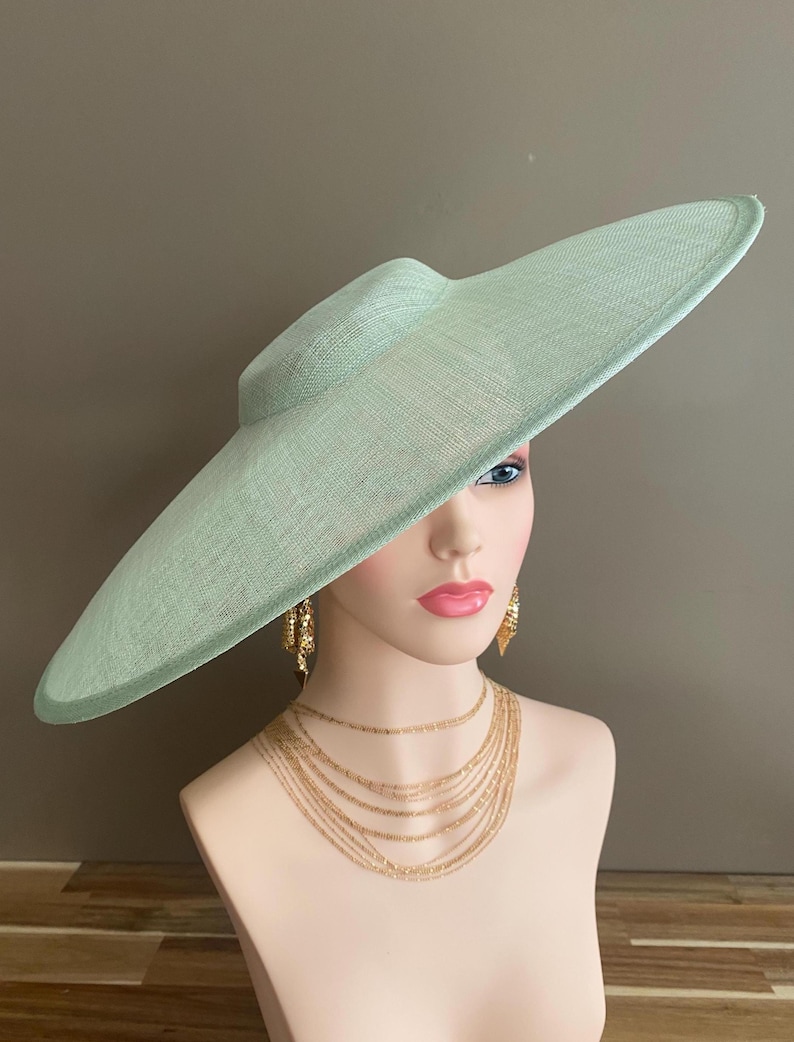 May include: A light green, wide-brimmed hat with a structured crown. The hat is displayed on a mannequin head, paired with gold-toned layered necklaces and earrings. The hat's brim has a subtle curve, and the material appears woven.