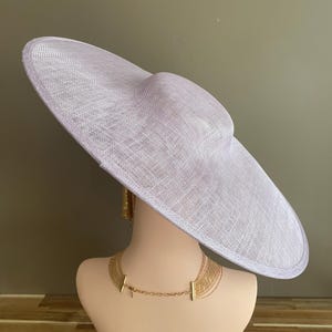 May include: A light purple, wide-brimmed hat with a rounded crown, displayed on a mannequin head. The hat has a woven texture and is paired with a gold necklace and earrings. The hat's brim is approximately 18 inches in diameter.