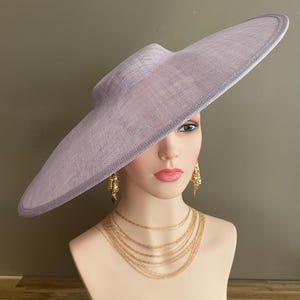 May include: A lavender-colored wide-brimmed hat with a structured crown. The hat is displayed on a mannequin head, paired with gold-toned layered necklaces and ornate earrings. The hat's brim extends outward, creating a dramatic silhouette.