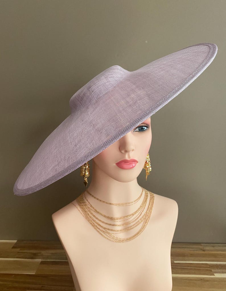 May include: A lavender-colored wide-brimmed hat with a structured crown. The hat is displayed on a mannequin head, paired with gold-toned layered necklaces and ornate earrings. The hat's brim extends outward, creating a dramatic silhouette.
