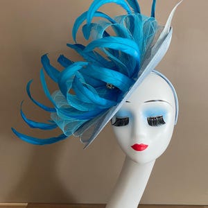 May include: A light blue hat with a large blue feather embellishment. The hat is worn on a white mannequin head.