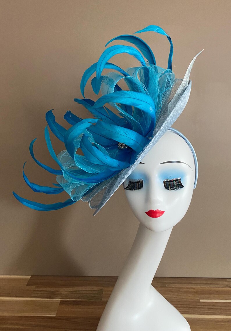 May include: A light blue hat with a large blue feather embellishment. The hat is worn on a white mannequin head.