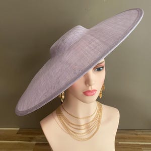 May include: A lavender-colored wide-brimmed hat with a structured crown. The hat is displayed on a mannequin head, paired with gold-toned layered necklaces and earrings. The hat's brim has a subtle, darker edge.
