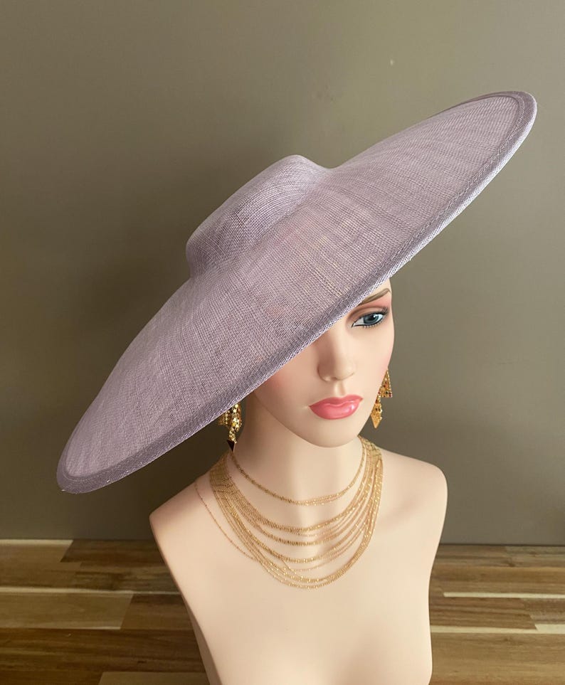 May include: A lavender-colored wide-brimmed hat with a structured crown. The hat is displayed on a mannequin head, paired with gold-toned layered necklaces and earrings. The hat's brim has a subtle, darker edge.