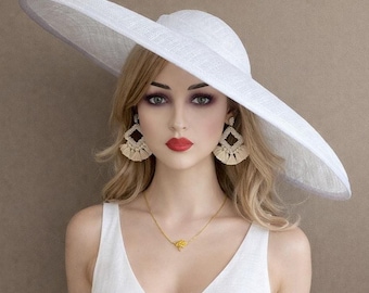 May include: A wide-brimmed white hat with a textured finish, paired with geometric diamond-shaped earrings featuring cream-coloured tassels and a delicate gold chain necklace with a small pendant, worn over a simple white sleeveless top.