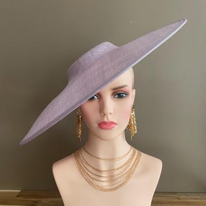 May include: A lavender-colored wide-brimmed hat with a structured design. The hat is displayed on a mannequin head, paired with gold-toned layered necklaces and dangling earrings. The hat's brim extends outward, creating a dramatic silhouette.