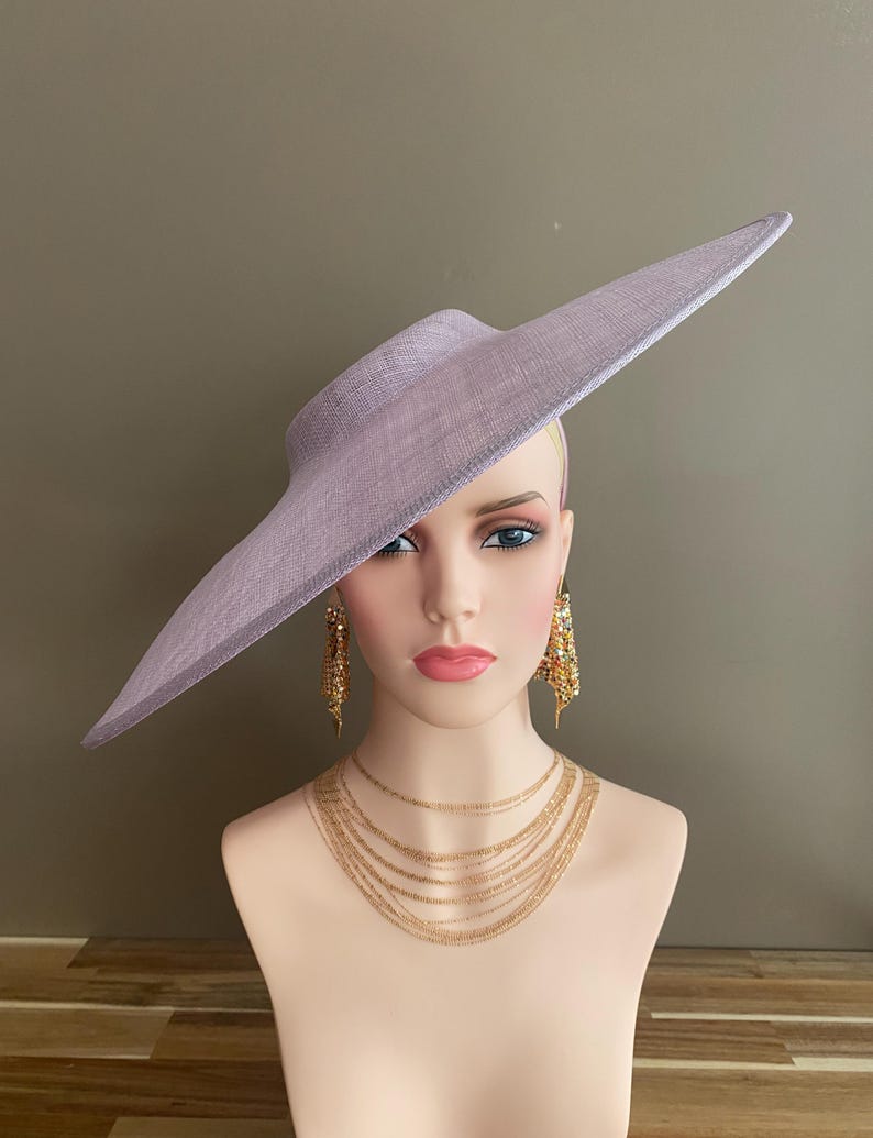 May include: A lavender-colored wide-brimmed hat with a structured design. The hat is displayed on a mannequin head, paired with gold-toned layered necklaces and dangling earrings. The hat's brim extends outward, creating a dramatic silhouette.