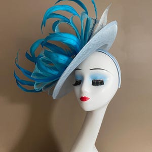 May include: A light blue fascinator with a large turquoise feather accent. The fascinator is worn on a white mannequin head.