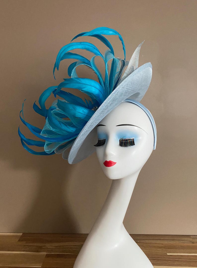 May include: A light blue fascinator with a large turquoise feather accent. The fascinator is worn on a white mannequin head.