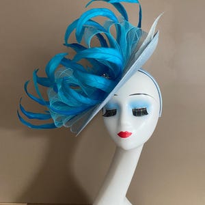 May include: A light blue fascinator with a large blue feather plume. The fascinator is worn on a mannequin's head.