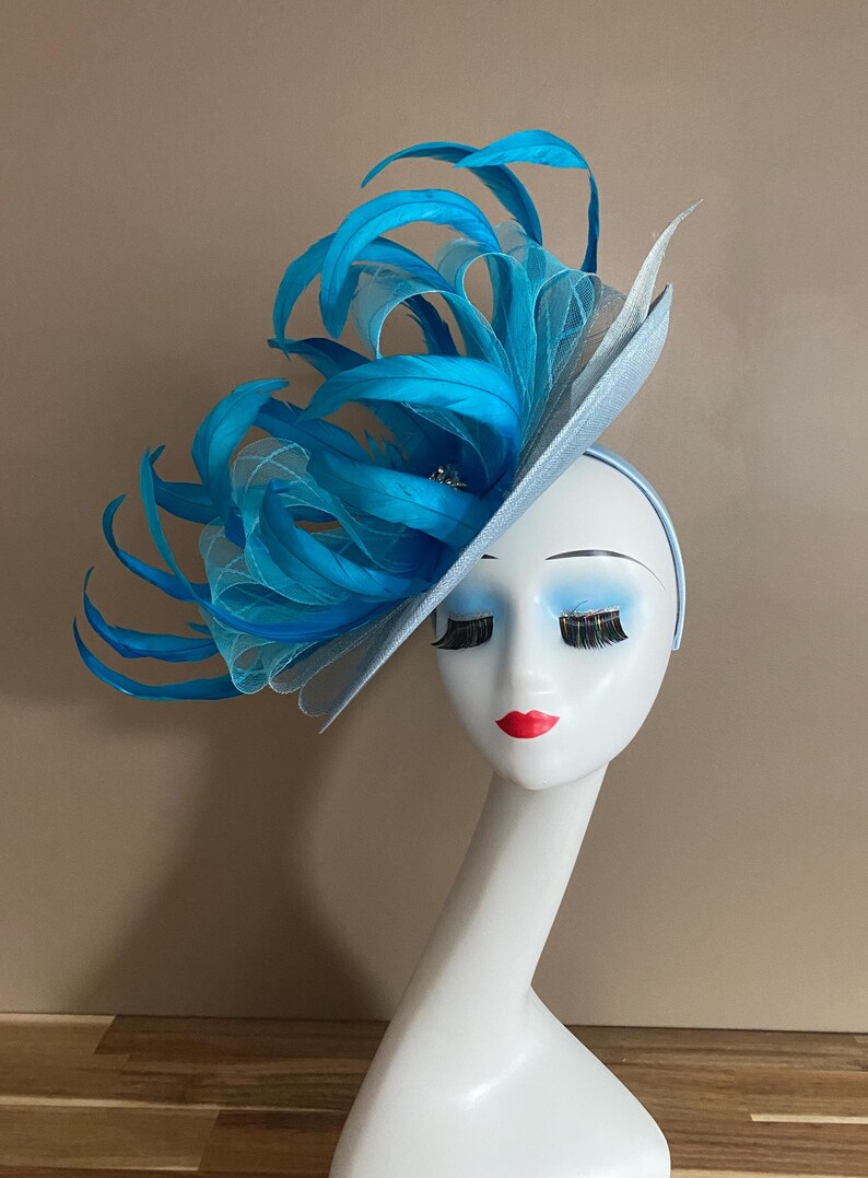 May include: A light blue fascinator with a large blue feather plume. The fascinator is worn on a mannequin's head.