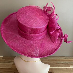 May include: A vibrant fuchsia hat with a wide brim and a cylindrical crown. The hat features a matching ribbon band and a decorative, swirling accent on the side. The hat is displayed on a mannequin head.