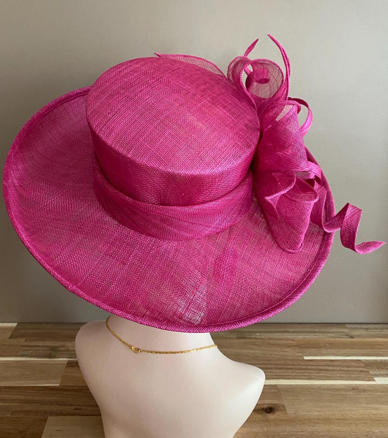 May include: A vibrant fuchsia hat with a wide brim and a cylindrical crown. The hat features a matching ribbon band and a decorative, swirling accent on the side. The hat is displayed on a mannequin head.