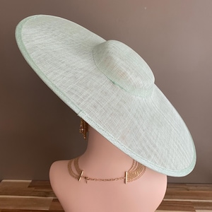 May include: A light green wide-brimmed hat with a rounded crown, displayed on a mannequin head. The hat has a woven texture and a delicate appearance. A gold necklace and earrings are also visible.