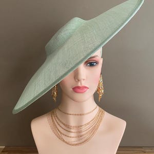 May include: A mint green wide-brimmed hat with a textured weave, displayed on a mannequin head. The mannequin also wears gold-toned layered necklaces and dangling earrings. The hat's brim extends dramatically to the side.