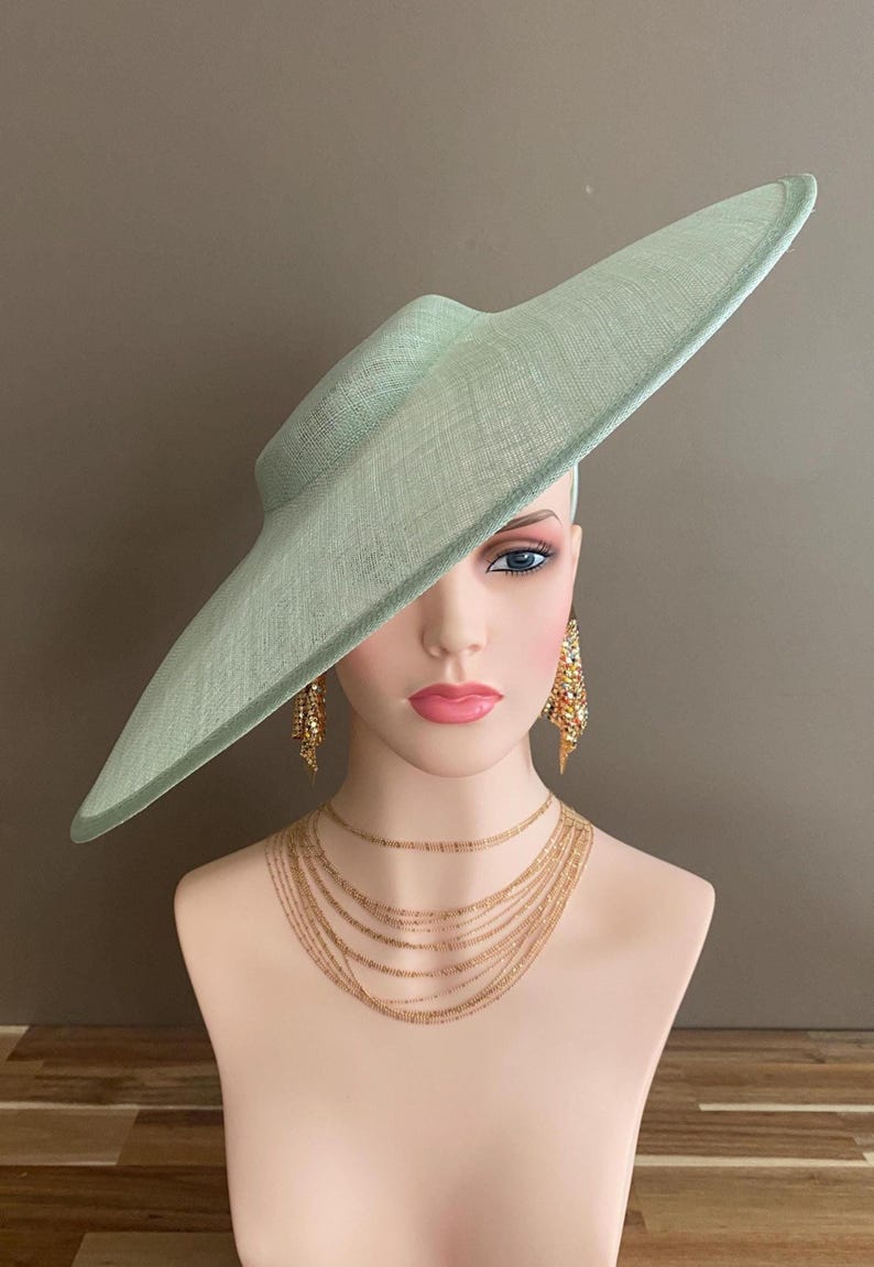 May include: A mint green wide-brimmed hat with a textured weave, displayed on a mannequin head. The mannequin also wears gold-toned layered necklaces and dangling earrings. The hat's brim extends dramatically to the side.