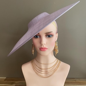 May include: A lavender-colored hat with a wide, asymmetrical brim. The hat is displayed on a mannequin head, paired with gold-toned layered necklaces and dangling earrings. The hat's brim extends to the side, creating a dramatic silhouette. The hat is a formal accessory.