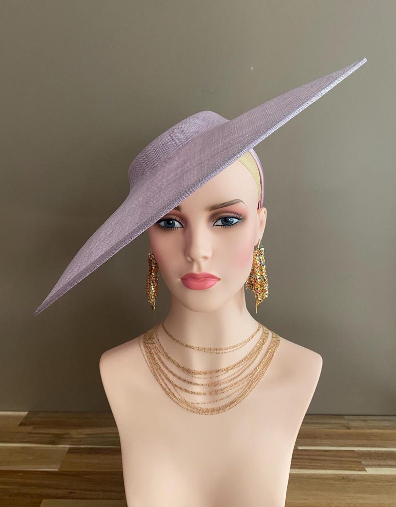 May include: A lavender-colored hat with a wide, asymmetrical brim. The hat is displayed on a mannequin head, paired with gold-toned layered necklaces and dangling earrings. The hat's brim extends to the side, creating a dramatic silhouette. The hat is a formal accessory.