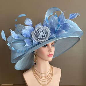 May include: A light blue wide-brimmed hat with a large, gray-blue flower and matching feathers. The hat is displayed on a mannequin head, paired with gold earrings and a layered gold necklace. The hat's brim is approximately 16 inches in diameter.