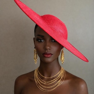 May include: A woman wearing a vibrant red hat with a wide, asymmetrical brim. She is adorned with gold earrings and a layered gold necklace. The hat is the focal point, complemented by the gold jewelry and a black strapless top.