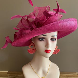 May include: A magenta-colored hat with a wide brim and decorative feathers. The hat is displayed on a mannequin head, paired with coral-colored earrings and a gold necklace. The hat's design includes a textured weave and layered fabric.