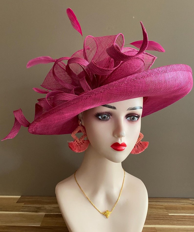 May include: A magenta-colored hat with a wide brim and decorative feathers. The hat is displayed on a mannequin head, paired with coral-colored earrings and a gold necklace. The hat's design includes a textured weave and layered fabric.