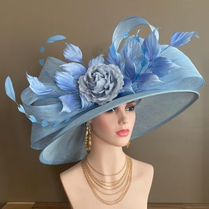 May include: A light blue wide-brimmed hat with a large gray rose and blue feather accents. The hat is displayed on a mannequin head, paired with gold earrings and a layered gold necklace. The hat's brim is approximately 15 inches in diameter.