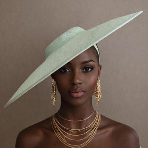 May include: A woman wearing a pale green wide-brimmed hat with a matching headband. She is also wearing multiple gold necklaces and dangling earrings. The background is a solid, neutral color.