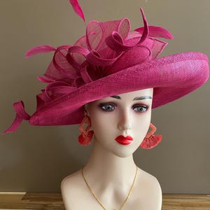May include: A magenta-colored hat with a wide brim and decorative mesh and feather accents. The hat is displayed on a mannequin head, paired with orange tassel earrings and a gold necklace.