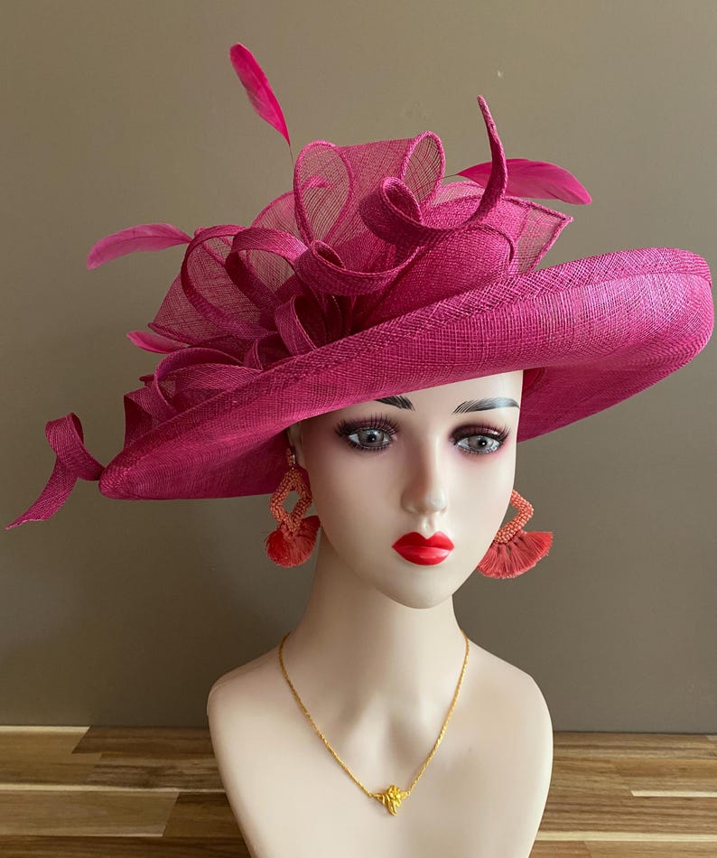 May include: A magenta-colored hat with a wide brim and decorative mesh and feather accents. The hat is displayed on a mannequin head, paired with orange tassel earrings and a gold necklace.