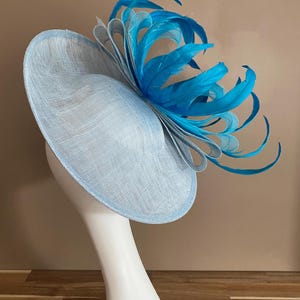 May include: A light blue hat with a large, bright turquoise feather accent. The hat is made of a woven material and has a wide brim.