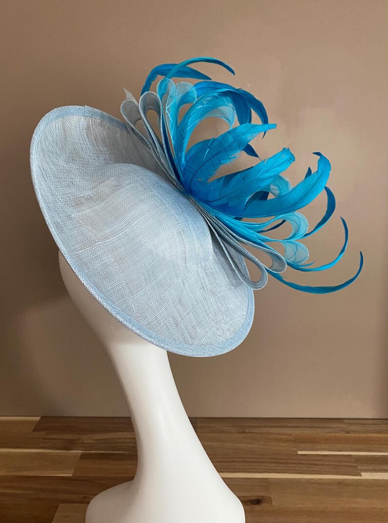 May include: A light blue hat with a large, bright turquoise feather accent. The hat is made of a woven material and has a wide brim.