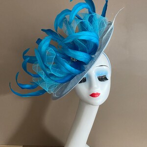 May include: A light blue hat with a large turquoise feather embellishment. The hat is made of a delicate, sheer fabric and is perfect for a special occasion.