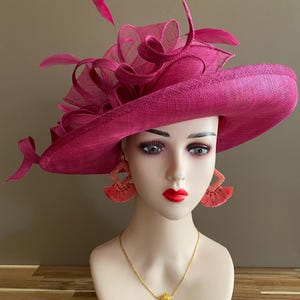 May include: A magenta-colored wide-brimmed hat with decorative fabric and feather accents. The hat is displayed on a mannequin head, paired with matching earrings and a gold necklace. The hat's brim measures approximately 15 inches in diameter.