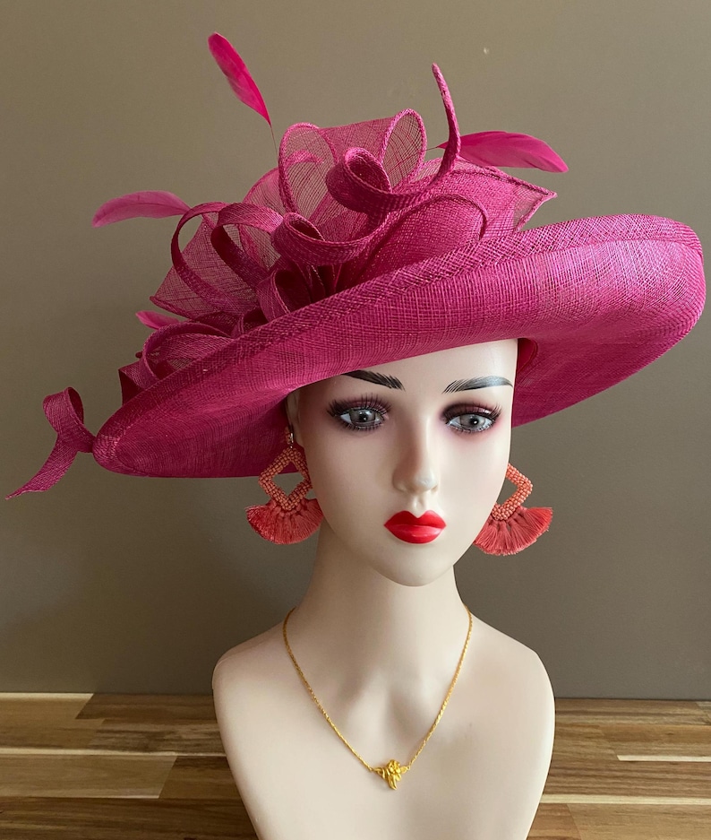 May include: A magenta-colored wide-brimmed hat with decorative fabric and feather accents. The hat is displayed on a mannequin head, paired with matching earrings and a gold necklace. The hat's brim measures approximately 15 inches in diameter.