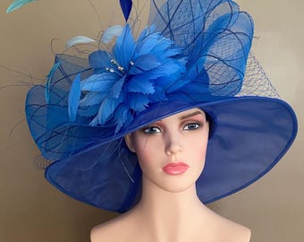May include: A vibrant blue hat with a wide brim, adorned with a large, layered flower and delicate feathers. The hat features a sheer, netted veil and is displayed on a mannequin head. A stylish accessory for special occasions.